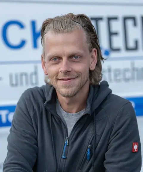 Techniker von LECKOTECH posiert lächelnd für ein Porträtfoto vor dem Firmenschild